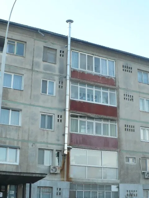 Coș de fum metalic pe fațada unui bloc, cu urme de rugină și de condens la bază