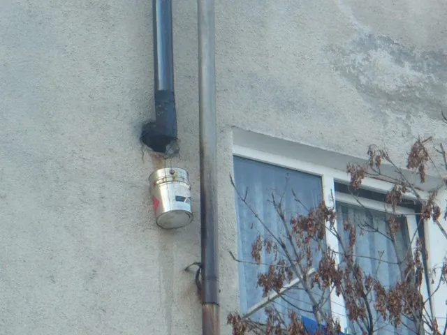 Găleată metalică agățată sub cotul unui coș de fum de pe fațada blocului, pentru colectarea condensului