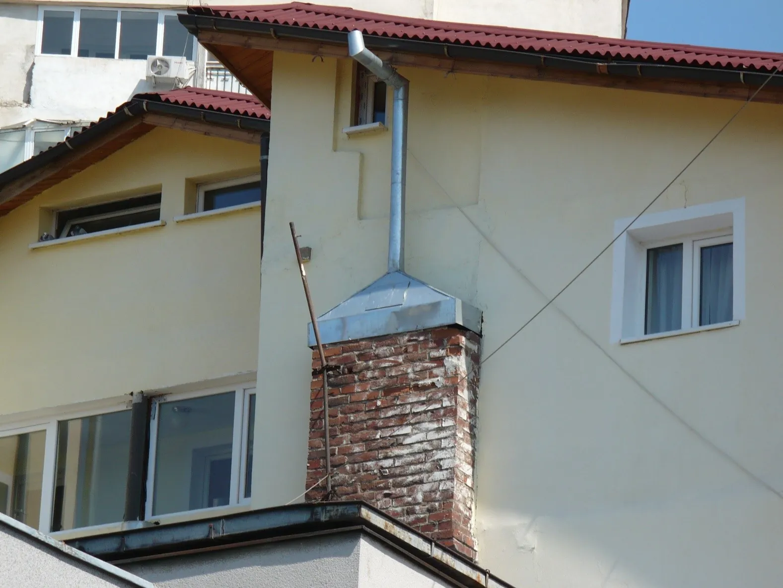 Coș de fum vechi din cărămidă, prelungit cu o țeavă metalică lipită de peretele clădirii noi, mai înalte