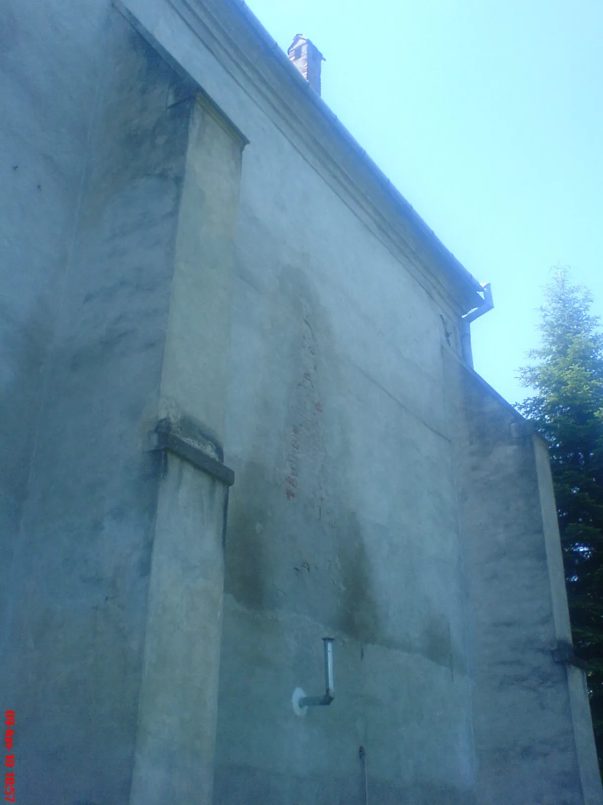 Peretele exterior al bisericii, cu coșul de fum din zidărie vizibil pe acoperiș și o țeavă metalică îndoită ieșind din zid, jos